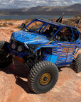 Blue FastLab off-road vehicle with rock slider steps driving on rocky terrain near a lake.