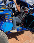 Man stepping into a blue off-road vehicle using rock slider steps on a dirt trail.